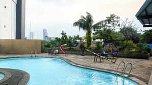 A swimming pool with a slide and swings, surrounded by greenery and buildings in the background.