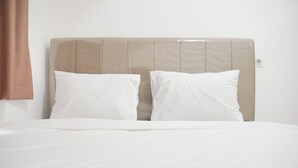 A bed with a beige headboard and white pillows, a brown curtain, and a white wall.