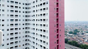 A high-rise building with a white and pink facade, multiple balconies, and a view of a cityscape with a highway in the foreground. - Comfort Designed 2Br Green Pramuka City Apartment (Jakarta)