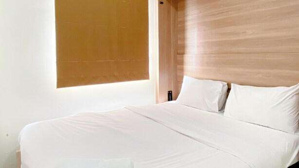 A modern bedroom with a large bed, a wooden headboard, and a window with blinds. - Comfort Designed 2Br Green Pramuka City Apartment (Jakarta)