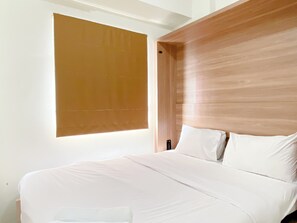 A modern bedroom with a large bed, a wooden headboard, and a window with blinds. - Comfort Designed 2Br Green Pramuka City Apartment (Jakarta)