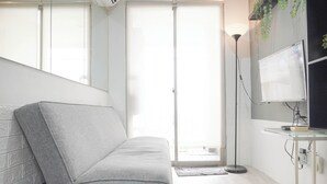 Una habitación moderna con un sofá gris, una lámpara de pie, un televisor de pantalla plana y un ventanal con persianas.