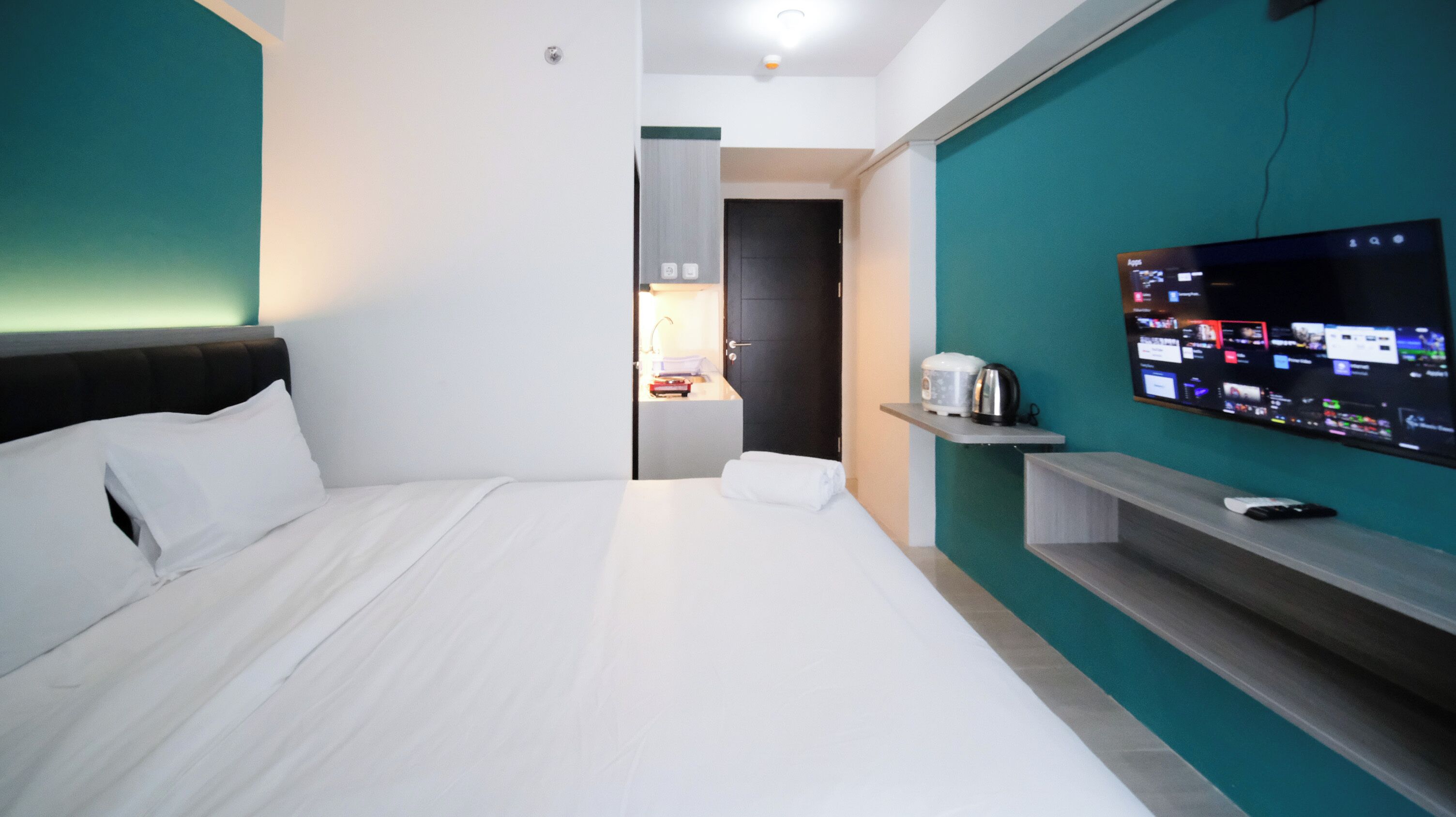 A modern hotel room with a large bed, a TV mounted on the wall, a small kitchenette, and a door leading to a bathroom.