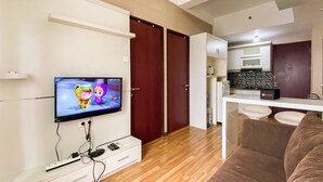 A modern living room with a flat-screen TV, wooden flooring, a brown sofa, and a kitchen area with white cabinets and a black tiled backsplash. - Signature 2Br At Apartment Tamansari Panoramic (Bandung)
