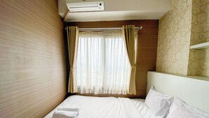 A small, single-bed room with a wooden headboard, a window with sheer curtains, and a wall-mounted air conditioner. - Signature 2Br At Apartment Tamansari Panoramic (Bandung)