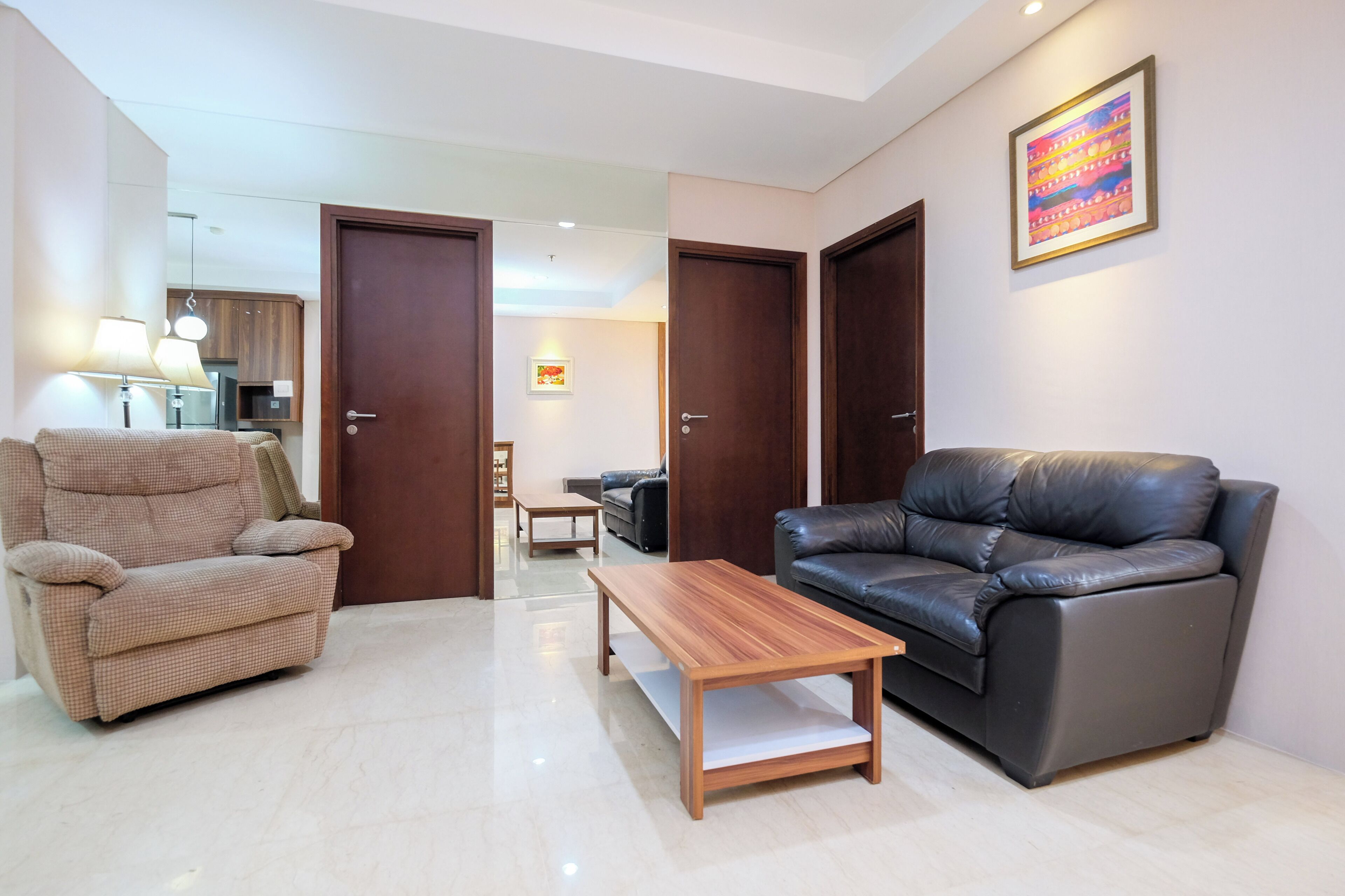 Una sala de estar con un sillón color beige, una mesa de centro de madera y un sofá de cuero negro. Hay dos puertas que dan a otras habitaciones, un cuadro en la pared y una lámpara a la izquierda.
