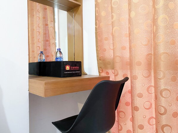 Une chambre d’hôtel avec une étagère en bois sur laquelle se trouve une boîte noire portant l’inscription « Travelio Property Management » et deux bouteilles d’eau. Une chaise noire est placée devant un miroir.