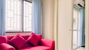 A bright room with a red sofa, a window with blue curtains, and a door leading to another room. - Good Deal And Homey Studio At Royal Sentul Park Apartment (Bogor)