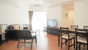 A modern living room with a black sofa, a wooden dining table with chairs, a flat-screen TV, and a large window with curtains. - Good Place 2Br Apartment At Sudirman Tower Condominium (Jakarta)