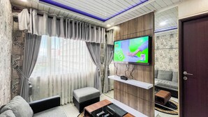 A living room with a TV mounted on a wooden wall, a grey sofa, a small ottoman, and a coffee table. - Cozy 2Br At Royal Makassar Apartment (Makassar)