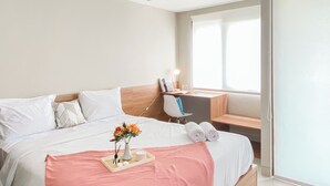 Une chambre d’hôtel comprenant un lit, un bureau, une chaise, une fenêtre et une table de chevet avec une lampe.
