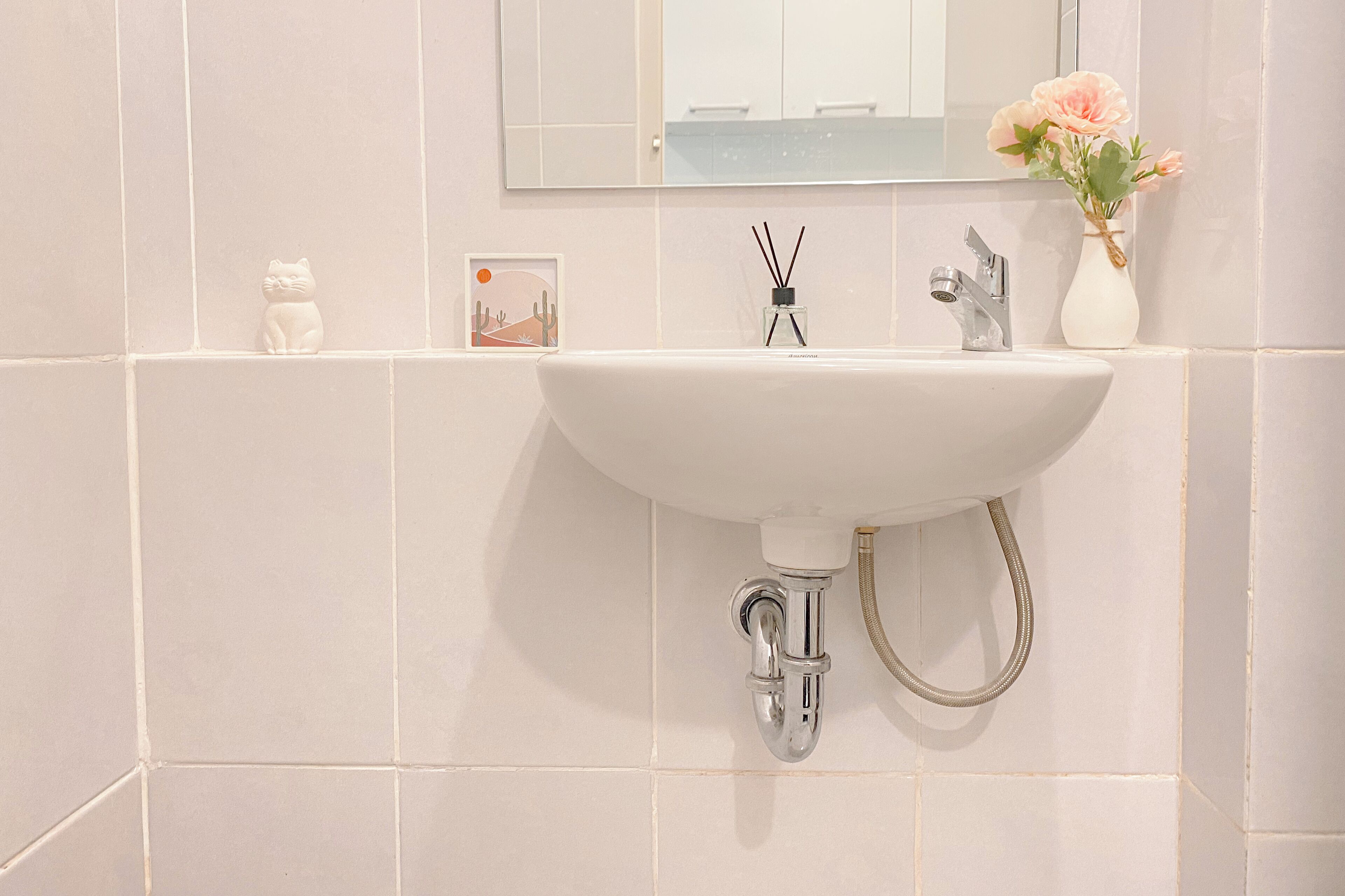 A bathroom with a white sink, a mirror, a small decorative cat, a picture frame, a vase with flowers, and a bottle of reed diffuser.