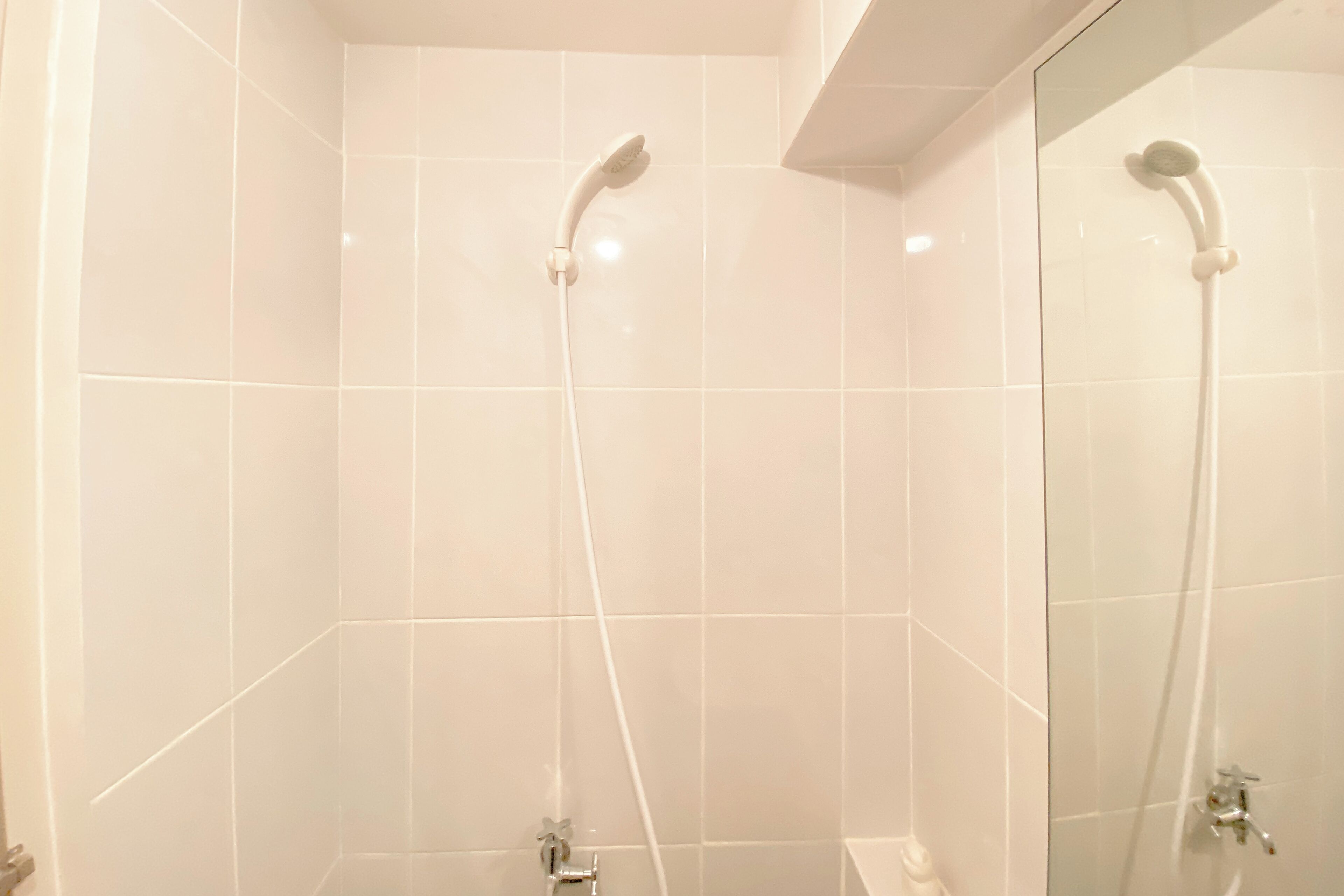 A bathroom with white tiles, a shower with a handheld showerhead, and a mirror.