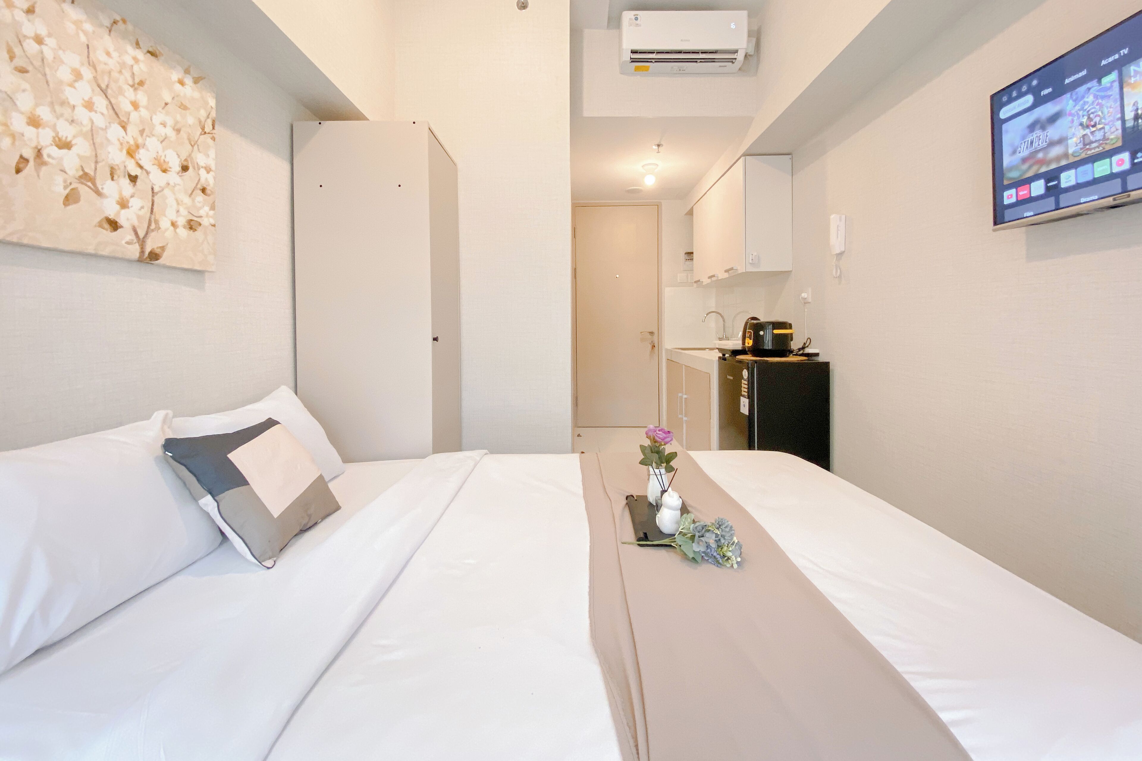 A hotel room with a bed, a small table with a vase and flowers, a television on the wall, and a kitchen area in the background.
