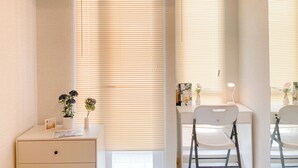 Habitación con escritorio, silla y un pequeño armario. Hay persianas en las ventanas y una lámpara sobre el escritorio.