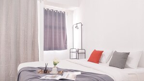 A neatly made bed with a grey bedspread, white pillows, and a red accent pillow. A small table with a vase of flowers and a book is on the bed. - Homey And Cozy Studio At Serpong Garden Apartment (Cisauk)
