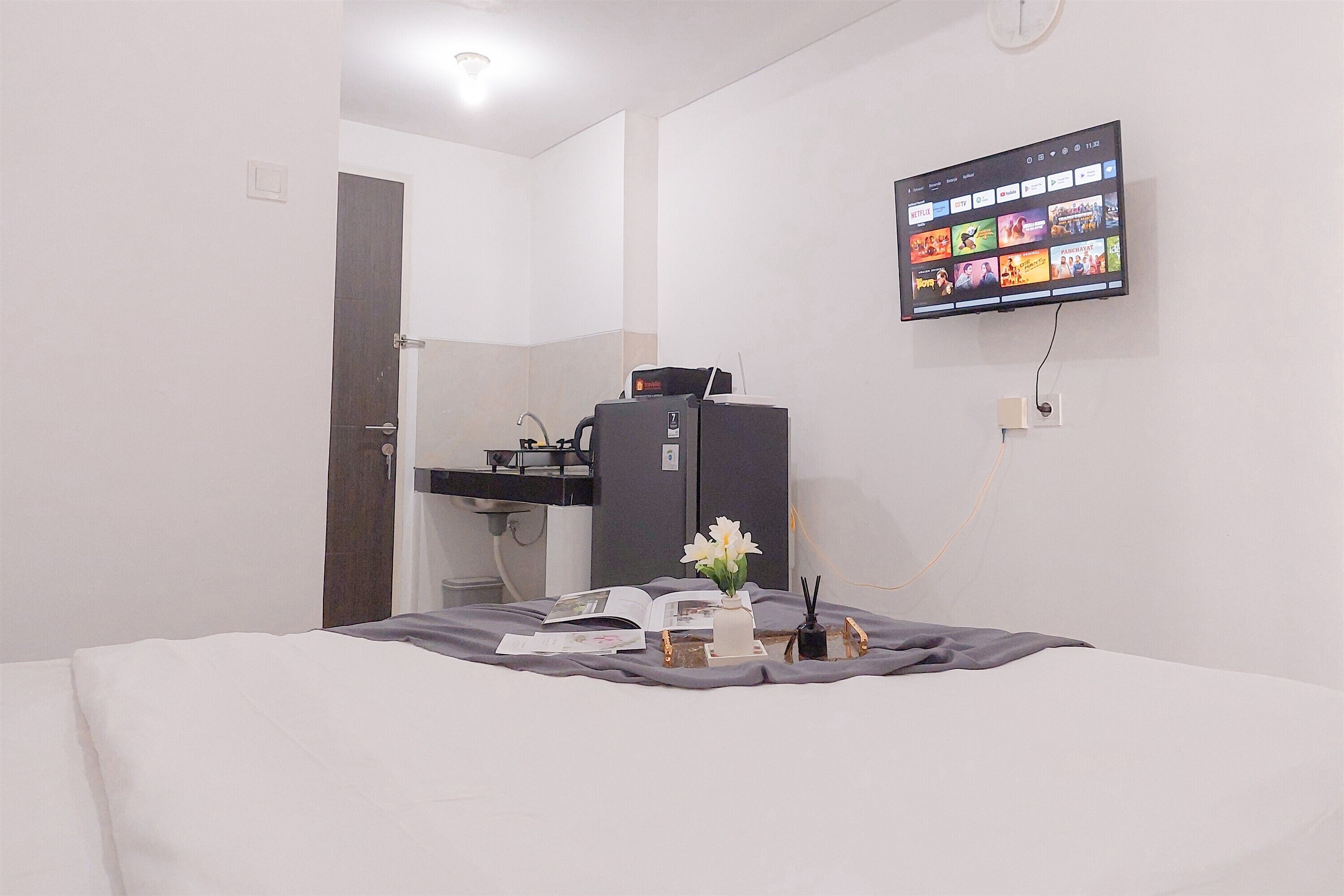A hotel room with a bed, a desk with a computer, a flat-screen TV mounted on the wall, and a small table with a vase of flowers.