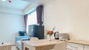 Une chambre d’hôtel compacte comprenant une table à manger en bois, une chaise blanche, une petite table d’appoint, un canapé, un téléviseur, une bouilloire et un vase décoratif avec des fleurs.