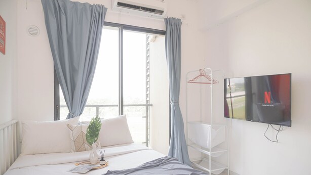 A bedroom with a bed, a TV, a window with curtains, and a plant. - Comfy And Best Deal Studio Sky House Bsd Apartment Near Aeon (Pagedangan)