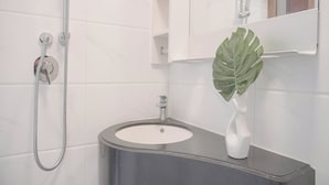 A bathroom with a white tiled wall, a black sink, a white vase with a green leaf, and a shower with a handheld showerhead. - Comfy And Best Deal Studio Sky House Bsd Apartment Near Aeon (Pagedangan)