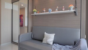 A modern living room with a grey sofa, a geometric patterned pillow, and a shelf with figurines and flowers. - Comfort And Good Place Studio At Sky House Bsd Apartment (Pagedangan)
