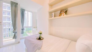 A modern hotel room with a large window, a bed with white bedding, a bedside table with a tray, and a shelf with decorative items. - Comfy Studio At Tokyo Riverside Pik 2 Apartment (Teluknaga)