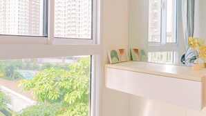 A room with a view of high-rise buildings, a window with a view of greenery, and a desk with decorative items. - Comfy Studio At Tokyo Riverside Pik 2 Apartment (Teluknaga)