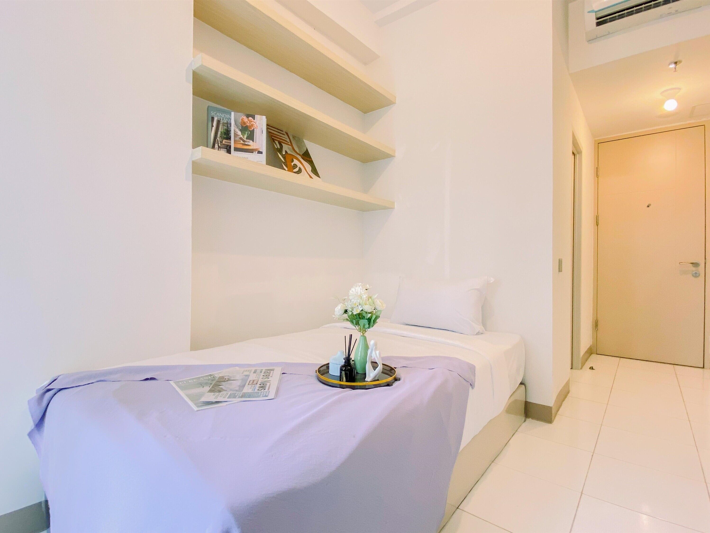 Un dormitorio pequeño y ordenado con una cama individual, una mesita de noche con un florero y una estantería con libros y adornos.