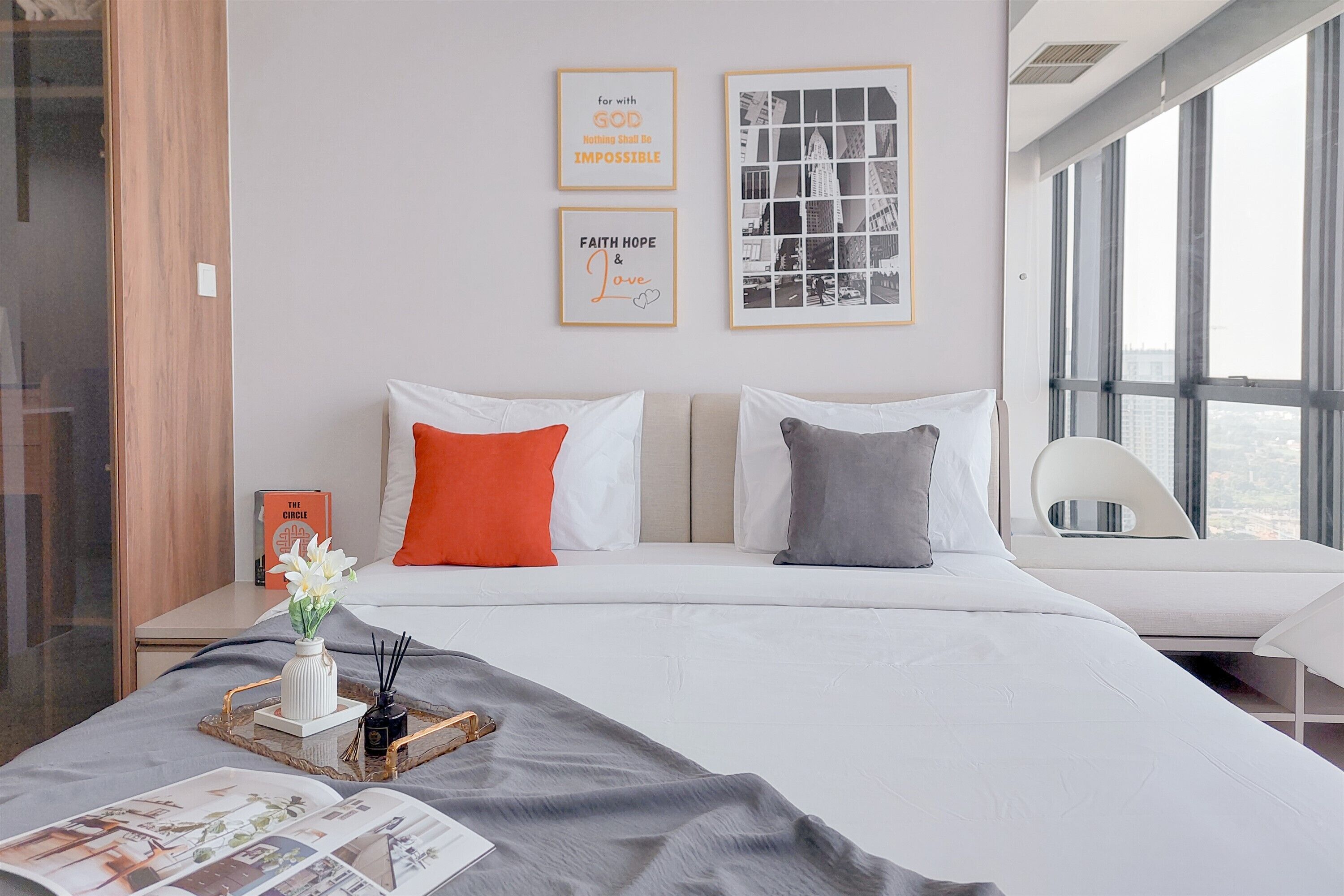 Una habitación de hotel moderna con una cama grande, dos almohadas, una mesita de noche con un libro, un jarrón con flores y cuadros en la pared.