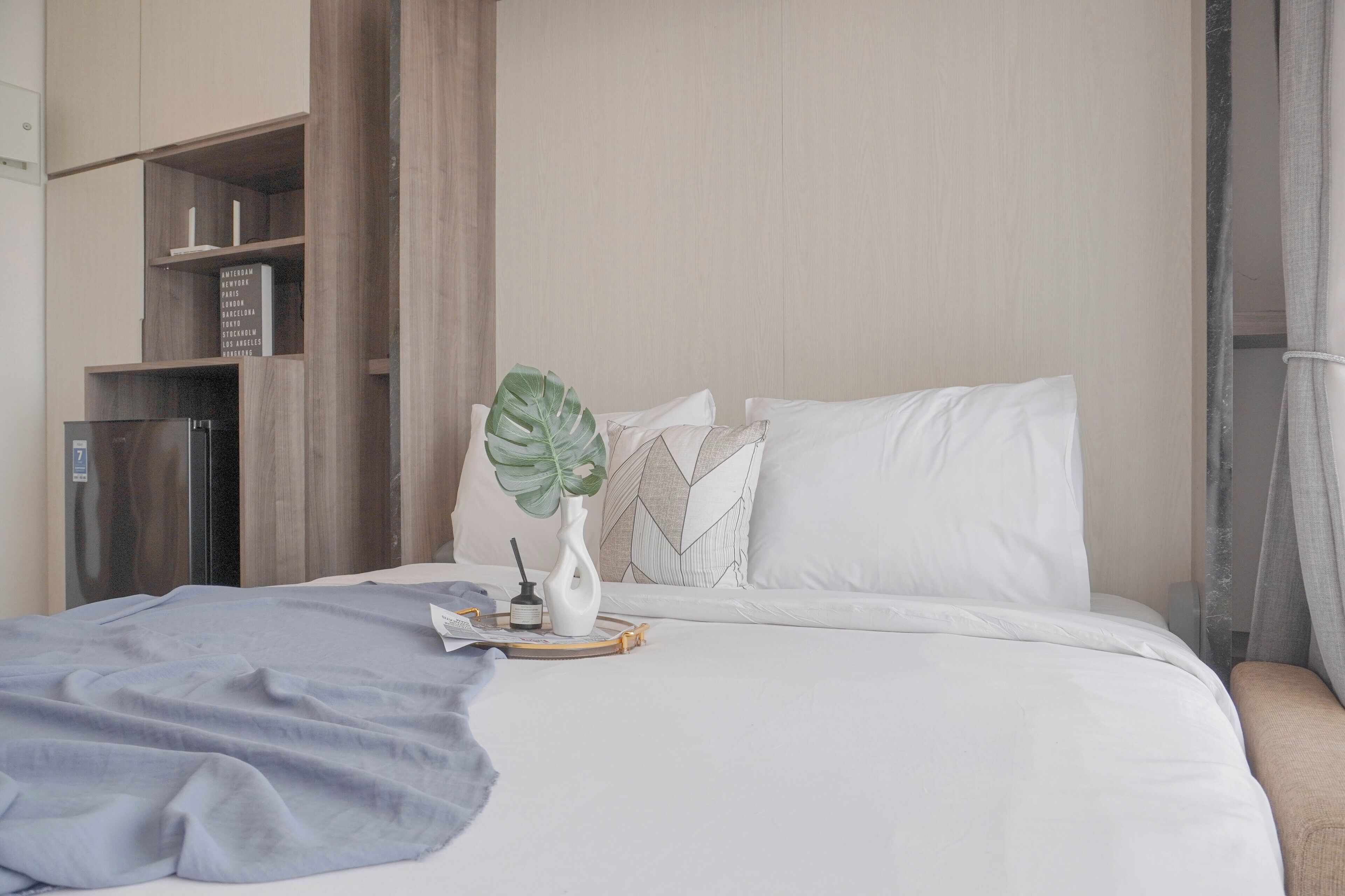 A neatly made bed with white linens, a gray throw blanket, and a decorative plant. There is a wooden shelf unit with books and a small table with a candle and a book.