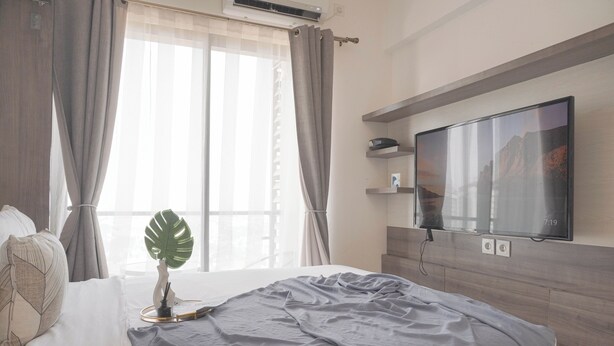 A hotel room with a bed, a TV mounted on the wall, a window with curtains, and a plant on the bedside table. - Best Choice And Cozy Studio Sky House Bsd Apartment (Pagedangan)