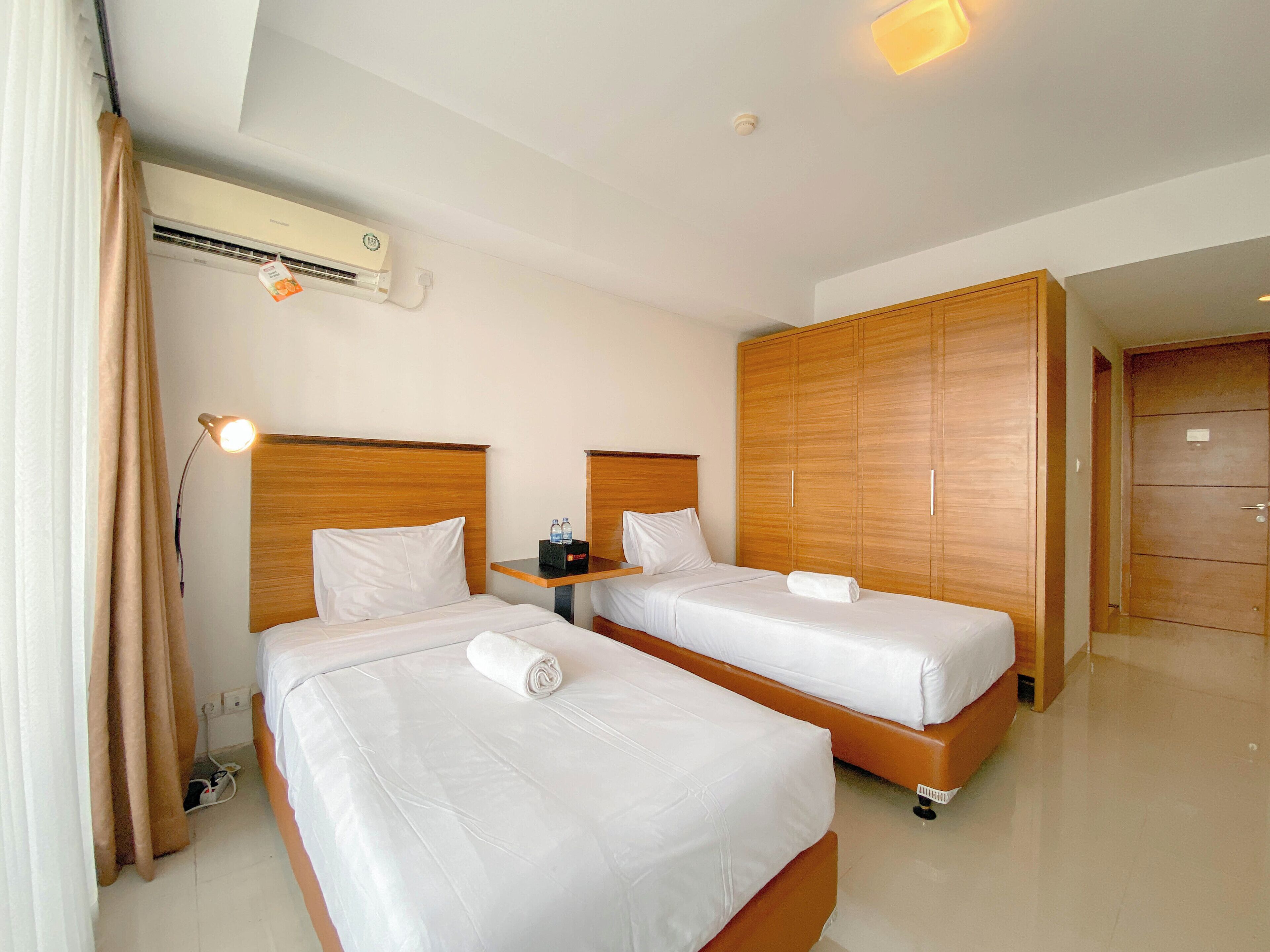 Habitación de hotel con dos camas, un armario de madera, una mesita de noche con lámpara y un aire acondicionado.