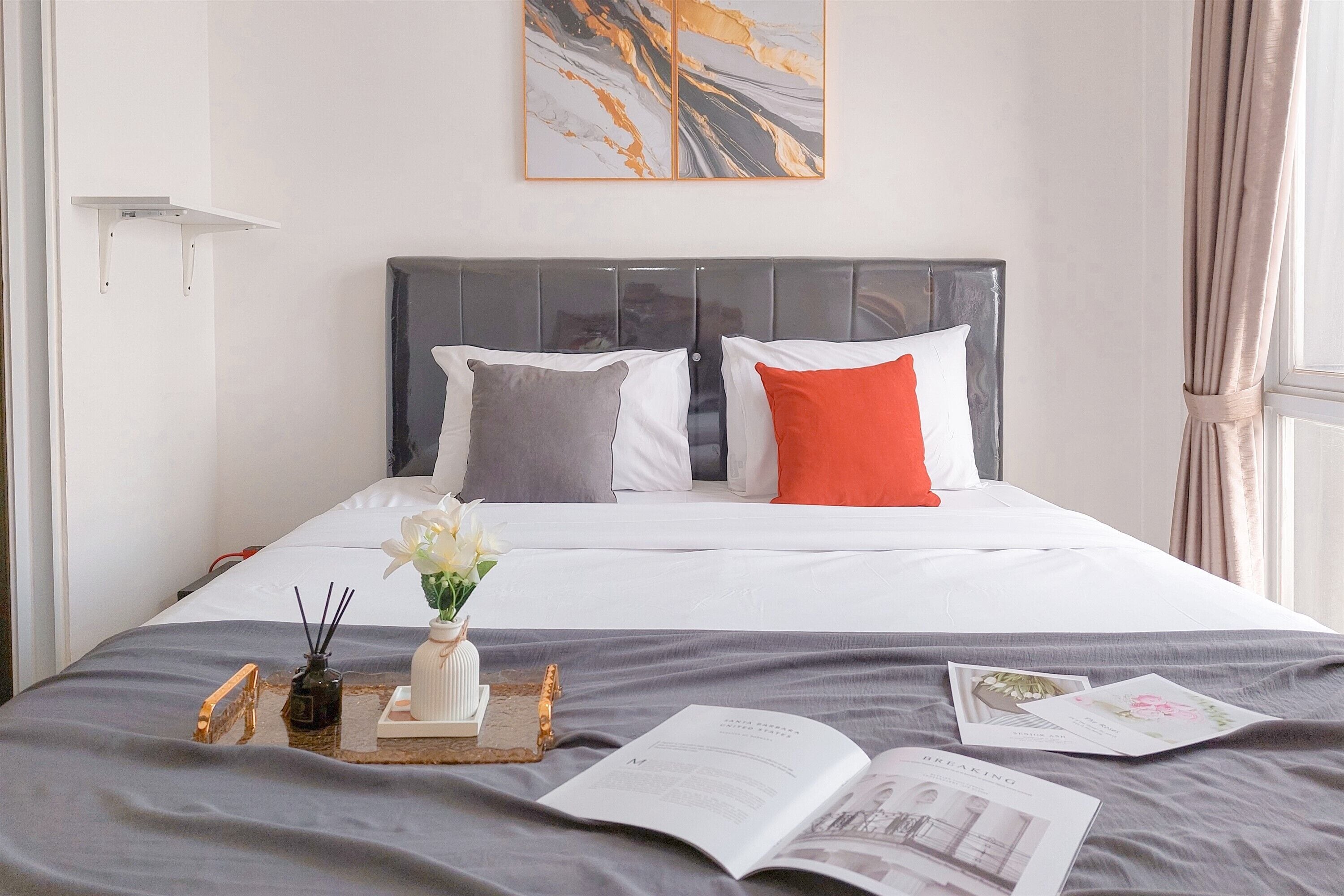 Un lit bien fait, avec une couette grise et blanche, un oreiller rouge et un oreiller gris. On peut voir un petit vase blanc avec des fleurs sur le lit.