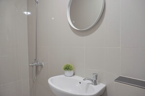 A bathroom with a white sink, a small potted plant, a round mirror, and a shower with a handheld showerhead. - Best Deal And Homey Studio Apartment Majestic Point Serpong (Gading Serpong)