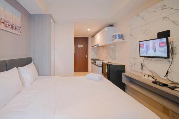 A hotel room with a bed, a television mounted on the wall, a small kitchen area, and a bathroom visible through an open door. - Compact And Comfortable Living Studio At Grand Sungkono Lagoon Apartment (Surabaya)