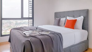 A modern bedroom with a large window, a grey upholstered headboard, and a neatly made bed with grey and white bedding. - Good Choice And Comfy 2Br Sky House Bsd Apartment (Pagedangan)
