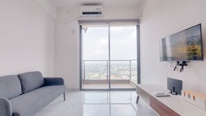 A modern living room with a grey sofa, a white TV stand, and a large window with a city view. - Good Choice And Comfy 2Br Sky House Bsd Apartment (Pagedangan)