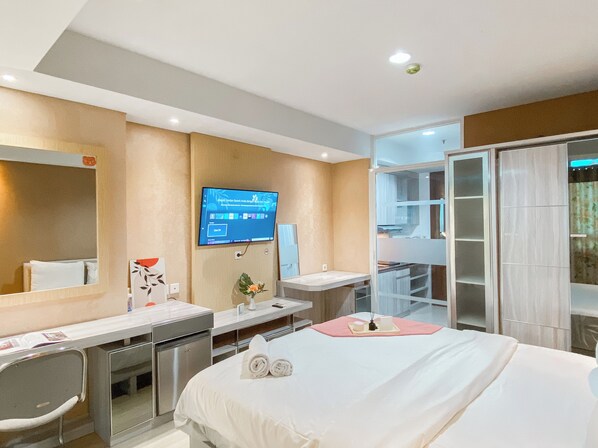 A hotel room with a bed, a desk, a TV mounted on the wall, a mirror, a wardrobe, and a bathroom visible through a glass door.