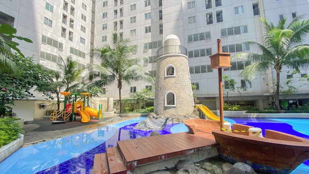 A swimming pool with a wooden boat and a stone tower, surrounded by palm trees and a playground. - Warm And Homey 2Br At Bassura City Apartment (Jakarta)