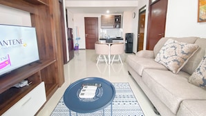 Uma sala de estar com televisão, sofá, mesa de centro e cozinha ao fundo.