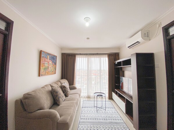 A living room with a sofa, a small table, a TV, and a painting on the wall. - Spacious And Minimalist 3Br At Gateway Pasteur Apartment (Cimahi)