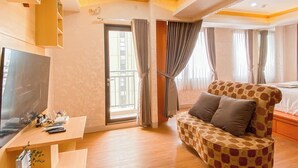 A hotel room with a sofa, a TV, a desk with a computer and other items, a balcony door, and curtains. - Good Place And Spacious Studio Transpark Cibubur Apartment (Depok)