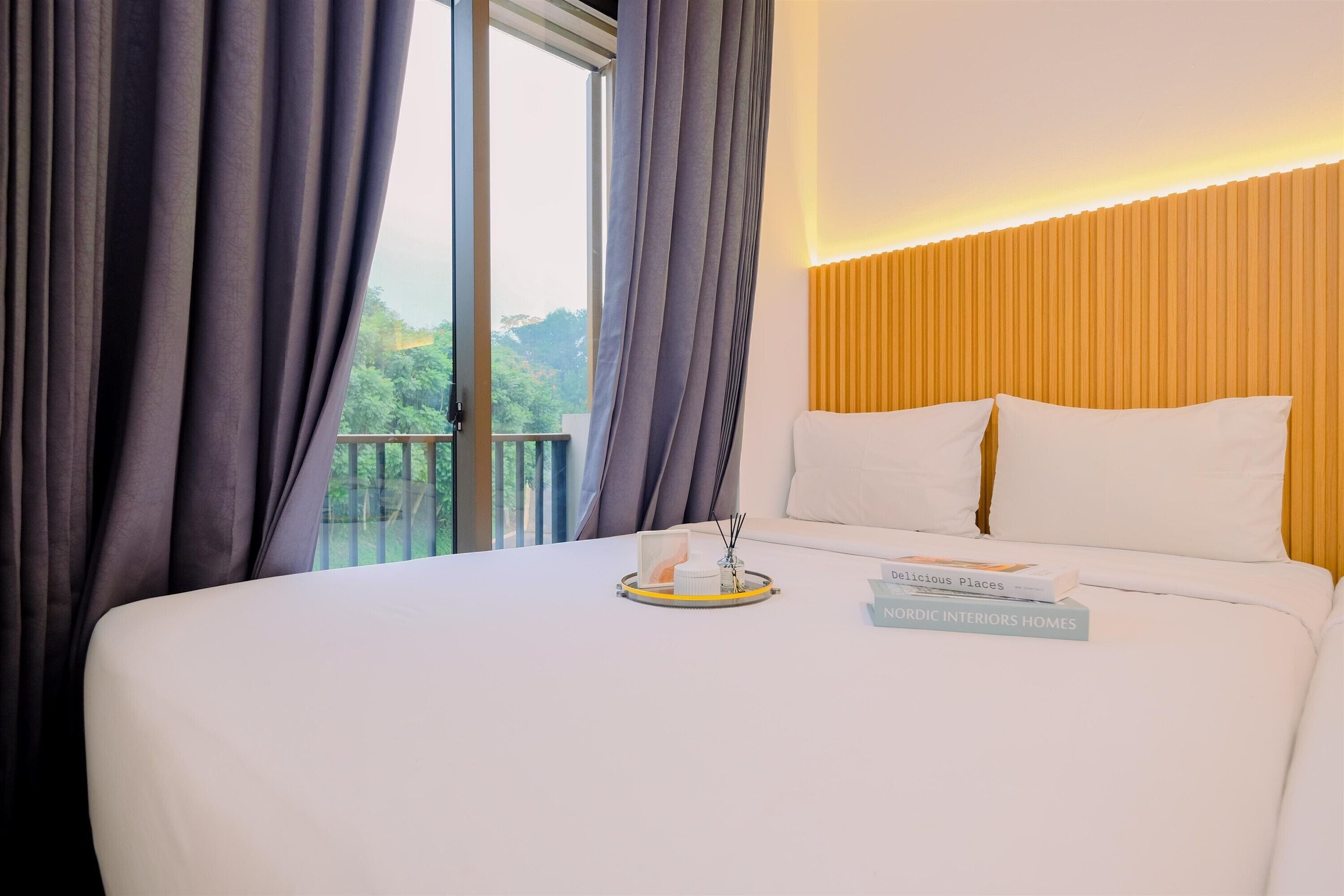 Une chambre d’hôtel avec un grand lit, une table de chevet sur laquelle se trouve un plateau avec des articles de toilette, et une vue sur la verdure par la fenêtre.