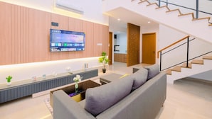 Una sala de estar moderna con un sofá gris, un televisor de pantalla plana montado en la pared y una escalera que conduce a un piso superior.