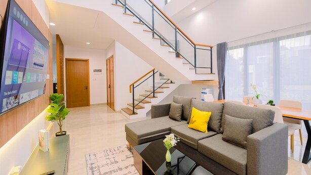 Una sala de estar moderna con un sofá gris, una mesa de centro, un televisor montado en la pared y una escalera que lleva a un piso superior.