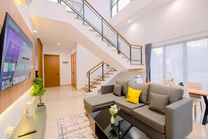 A modern living room with a grey sofa, a coffee table, a TV mounted on the wall, and a staircase leading to an upper floor. - Vimala Hills Villa Gadog Puncak 3Br For 12 Pax (Ciawi)