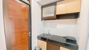 A compact kitchen with a wooden door, a sink, cabinets, and a trash bin. - Homey Designed 3Br At Gateway Pasteur Apartment (Cimahi)