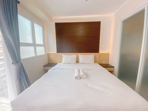 A hotel room with a large bed, two bedside tables, a wooden headboard, and a window with curtains. - Homey Designed 3Br At Gateway Pasteur Apartment (Cimahi)