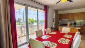 A dining area with a red tablecloth, white chairs, and a view of the outdoors through a sliding glass door.