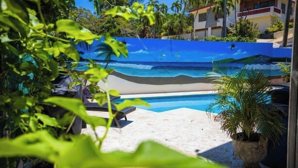 A pool area with a mural of a beach scene, surrounded by potted plants and a building in the background.